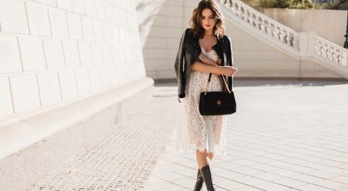 young stylish woman walking in street in fashionable outfit, holding purse, wearing black leather jacket and white lace dress, spring autumn style, posing, high leather boots